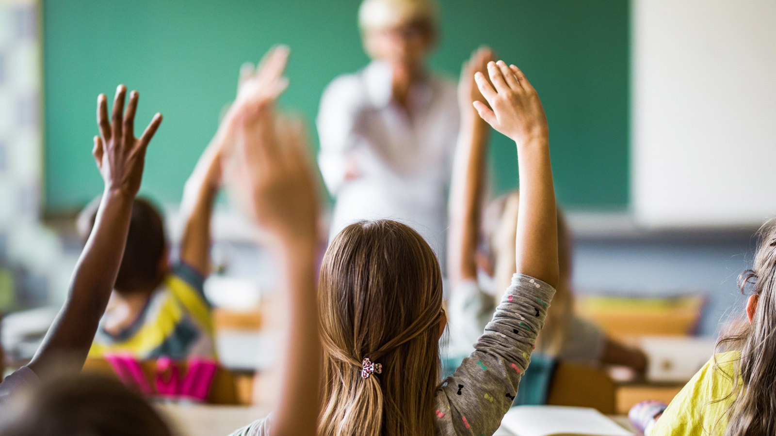 Schulkinder melden sich im Unterricht, im Hintergrund eine Lehrkraft an der Tafel