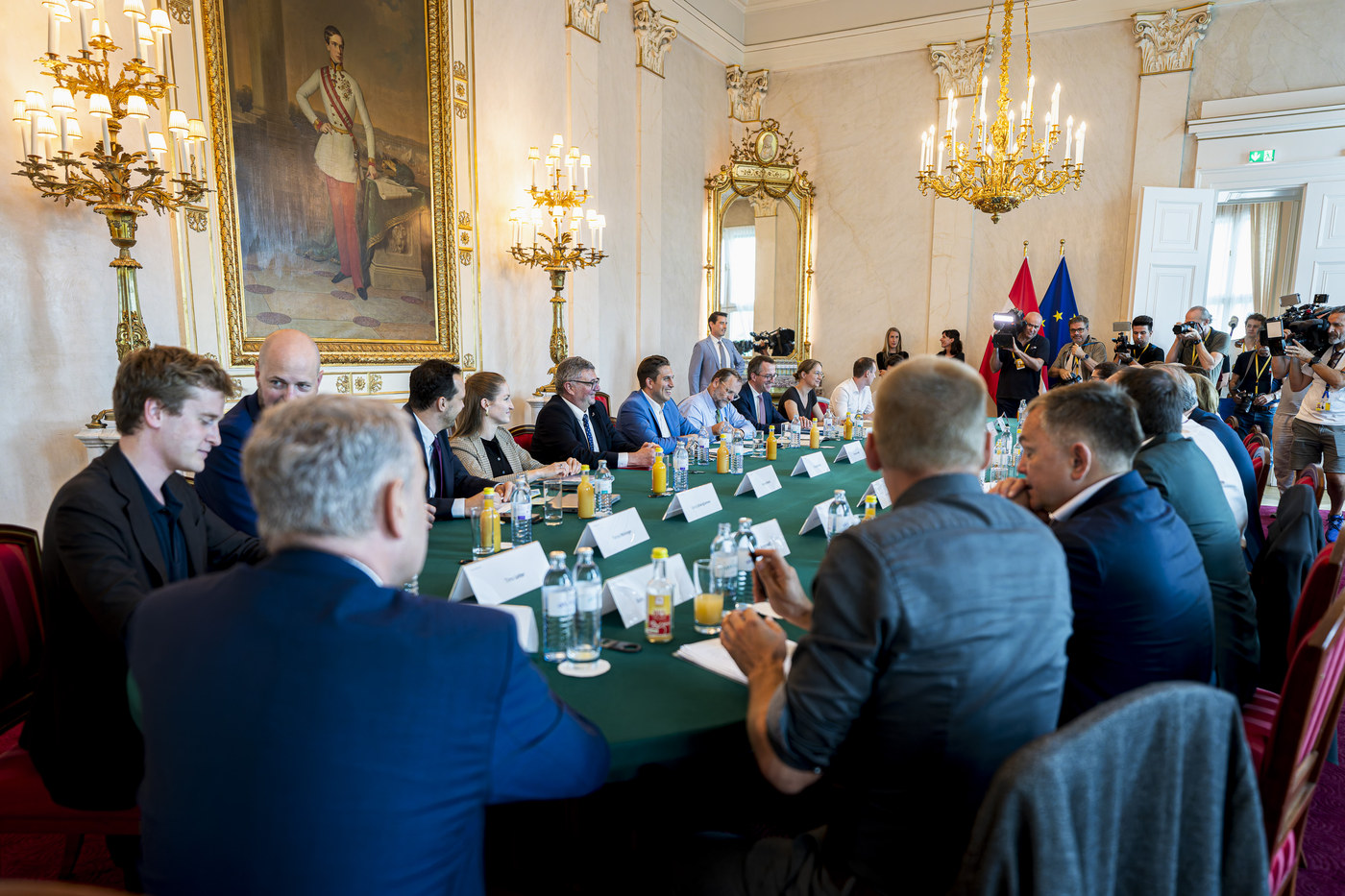 Verhandlungsrunde am grünen Tisch im Bundeskanzleramt, im Hintergrund Medienvertreterinnen und Medienvertreter